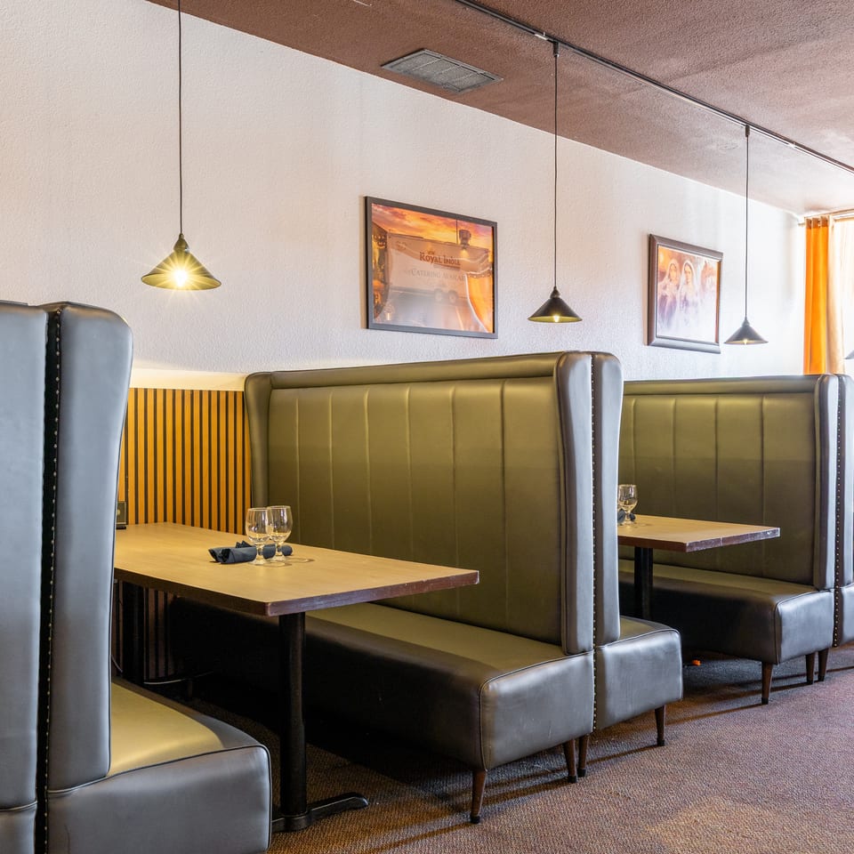 Elegant Indian restaurant dining room with leather booths, wooden tables, pendant lighting, framed artwork, and orange curtains.