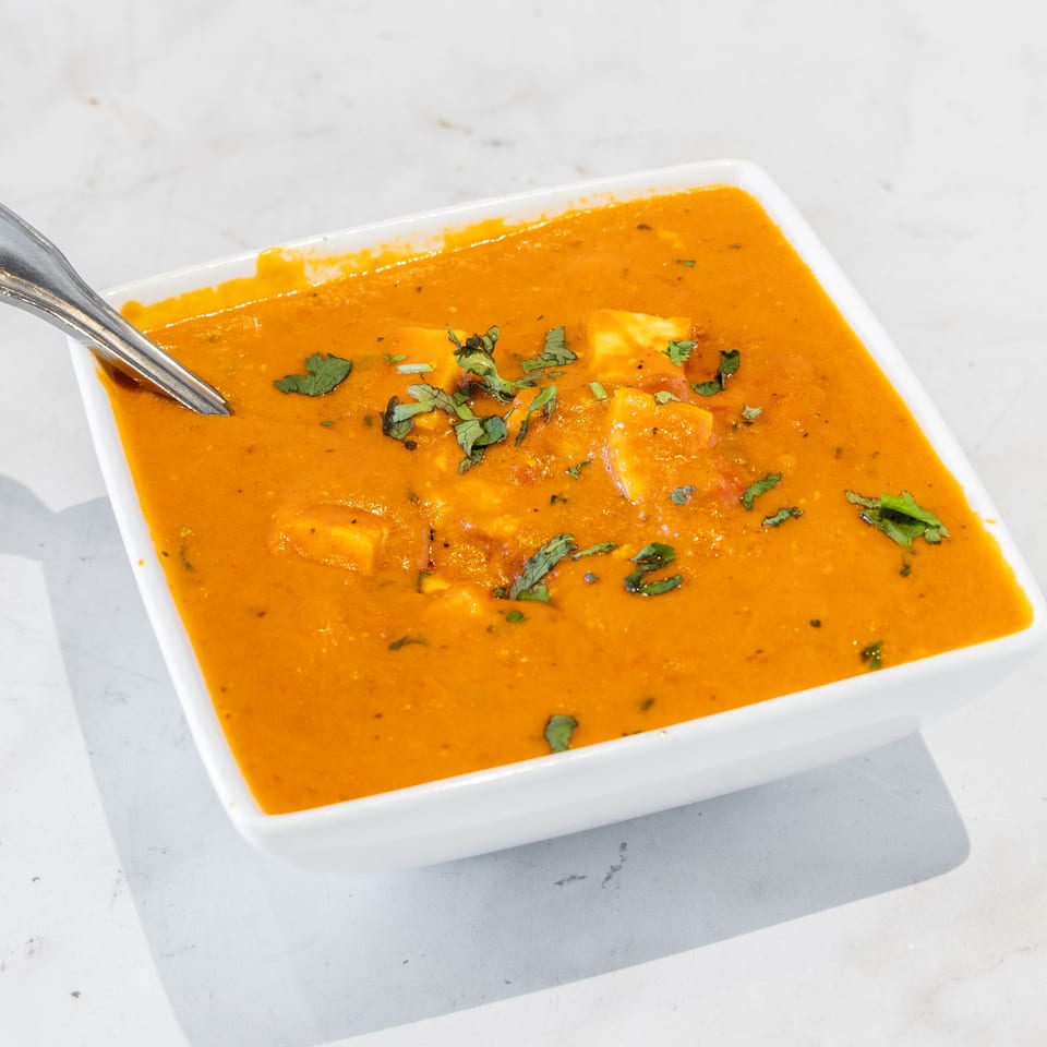 Square white dish of creamy orange curry with paneer cubes and cilantro garnish, served with a spoon.