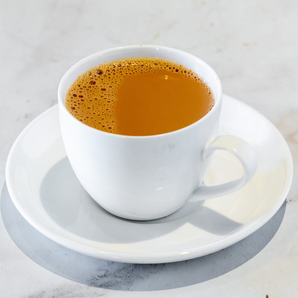 White cup of foamy coffee on a matching saucer atop a light-colored table.