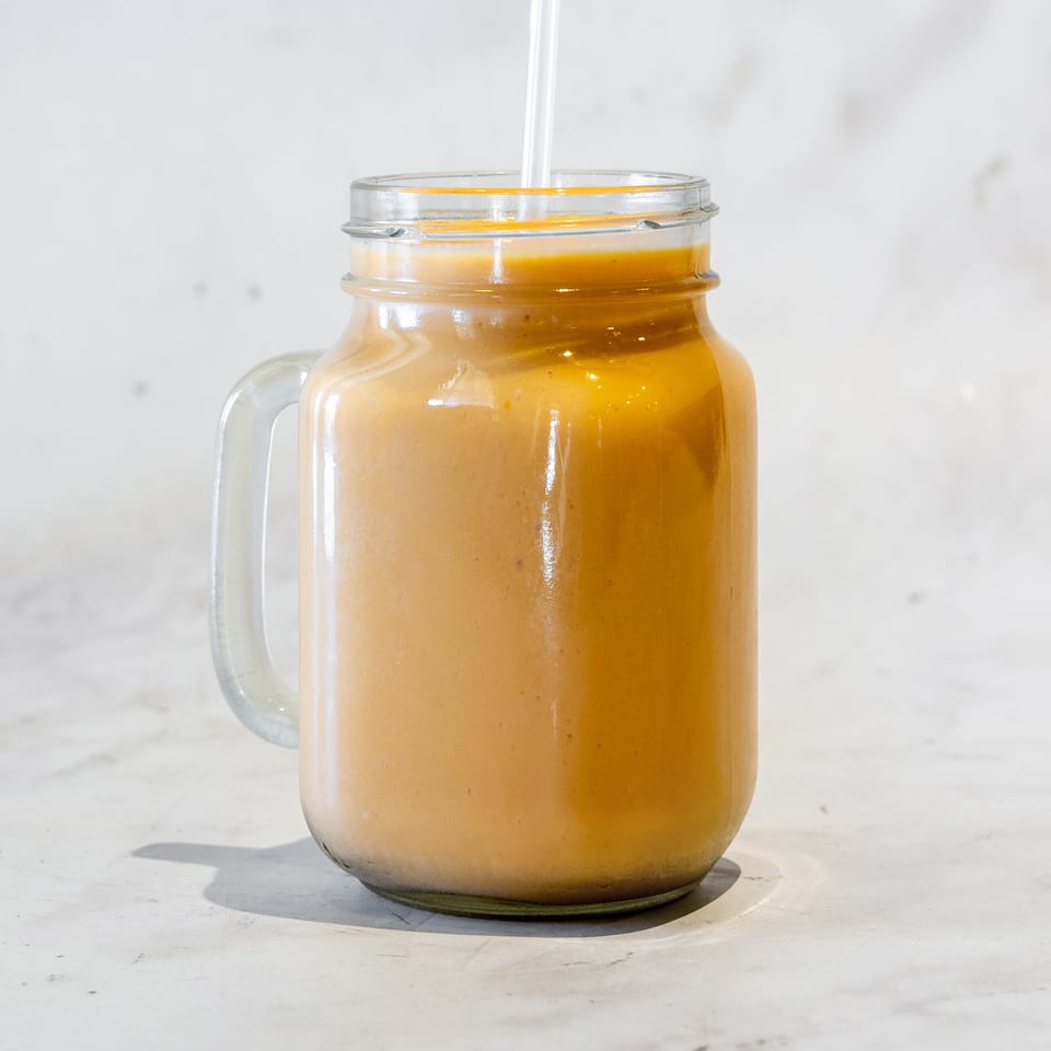 Golden-orange smoothie served in a clear glass mug with a straw on a light marble surface.