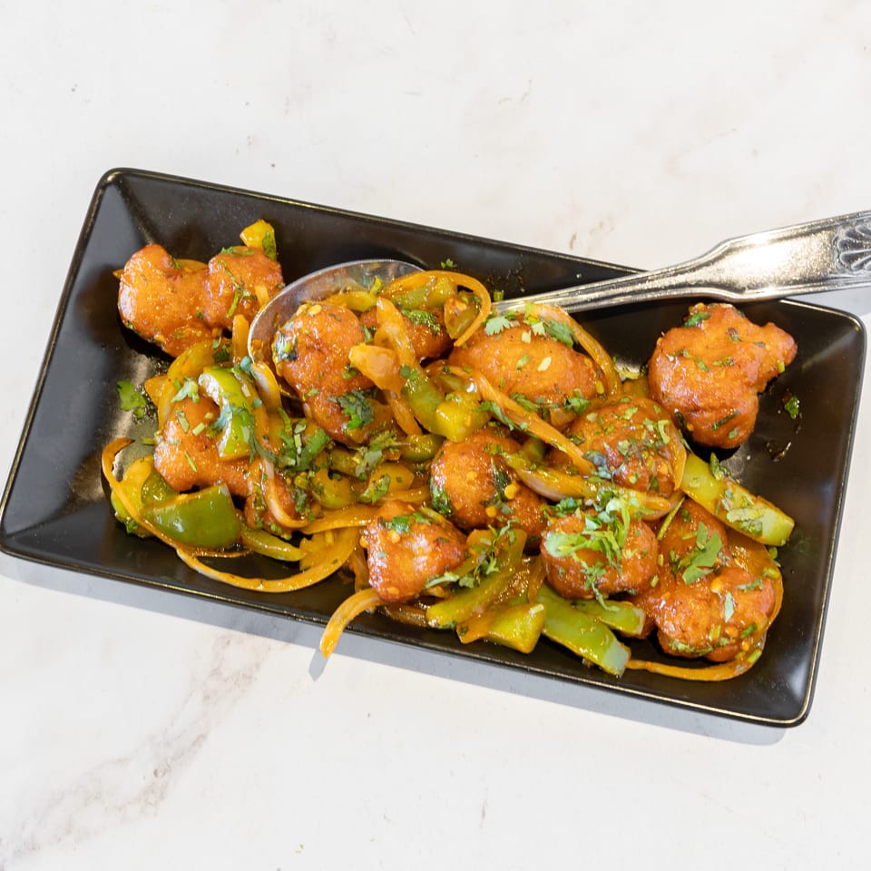 Spicy battered vegetable bites topped with sautéed peppers, onions, and cilantro on a black rectangular plate.