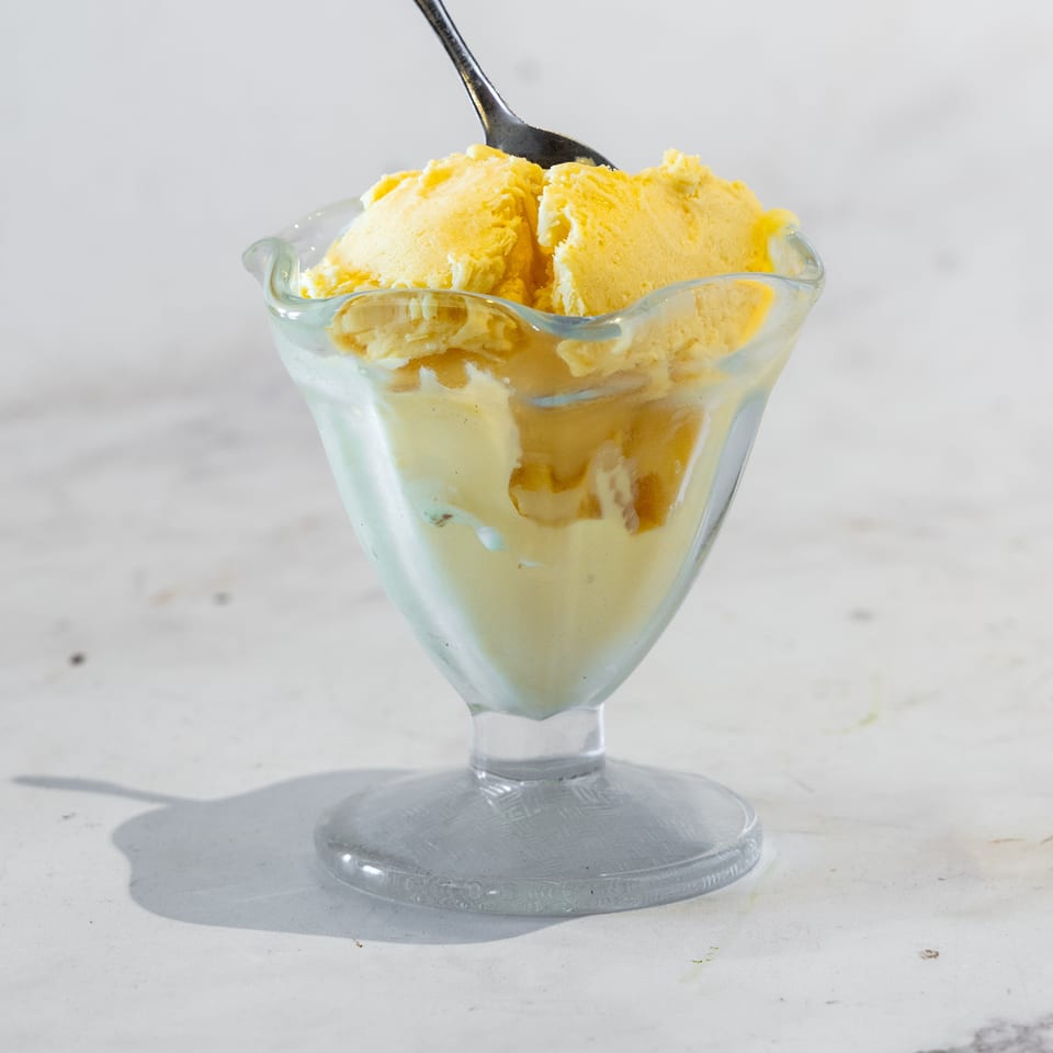 Scoops of yellow ice cream with caramel sauce in a glass dessert dish, topped with a spoon.