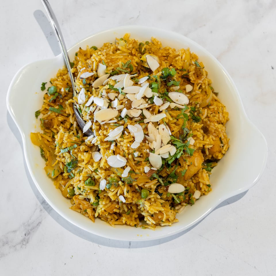 Bowl of golden spiced rice with peas, vegetables, almonds, raisins, and fresh cilantro, served with a spoon.