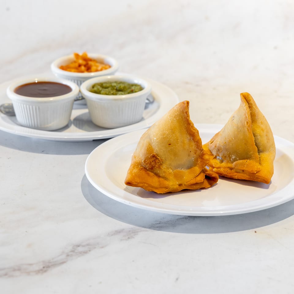 Golden fried samosas on a white plate with bowls of brown, green, and orange dipping sauces on a marble table.