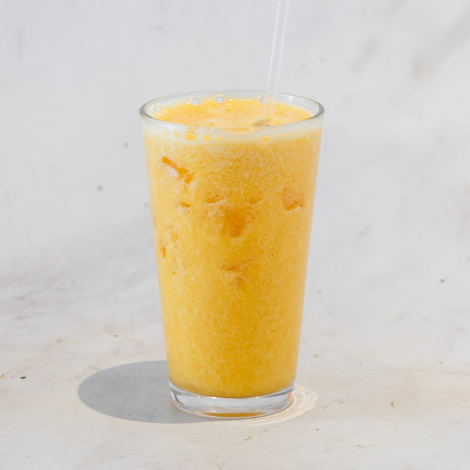 Tall glass of frothy orange juice with a straw on a sunlit white table.