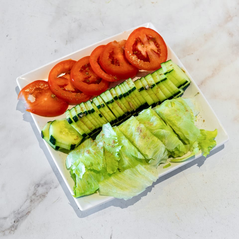 White plate of sliced tomatoes, cucumbers, and lettuce seasoned with herbs on a light marble surface.