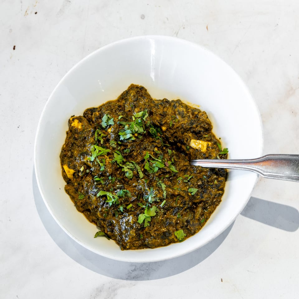 Bowl of dark green spinach curry with paneer cubes, garnished with fresh cilantro and served with a spoon.