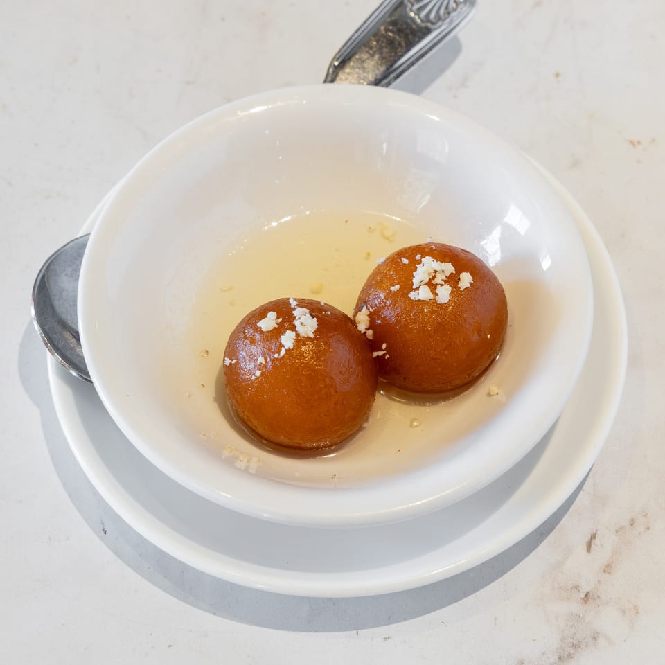 Two syrup-soaked gulab jamun topped with crumbled white garnish in a white bowl with spoon.