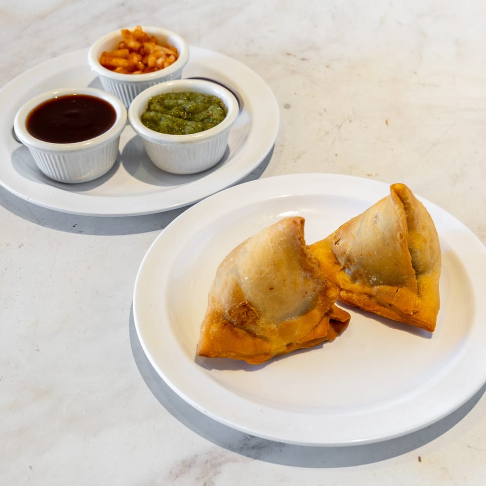Golden samosas on a white plate beside ramekins of dark, green, and orange dipping sauces.